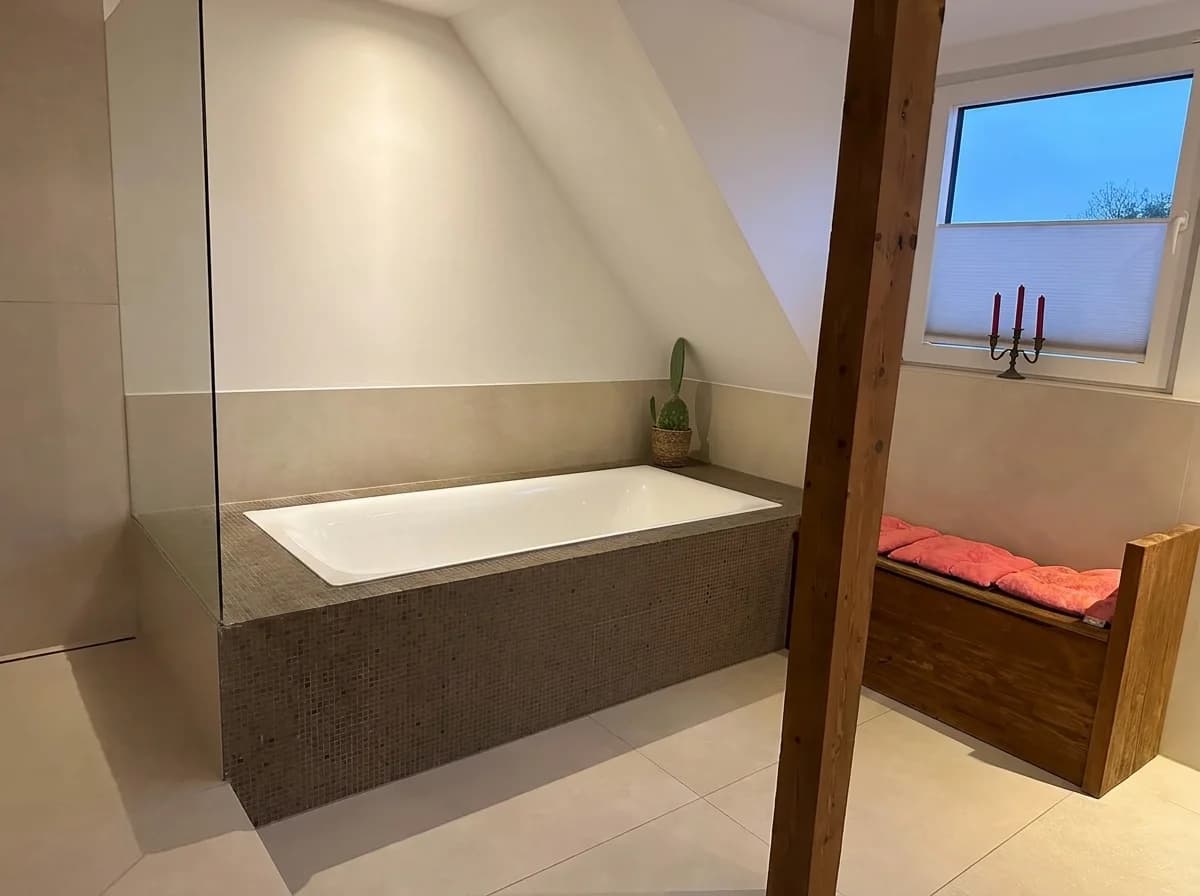 Badewanne mit Mosaikverkleidung unter der Dachschräge, daneben eine Sitztruhe aus Altholz